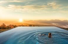 Person swimming in an infinity pool with panoramic view of mountain range and sunset.