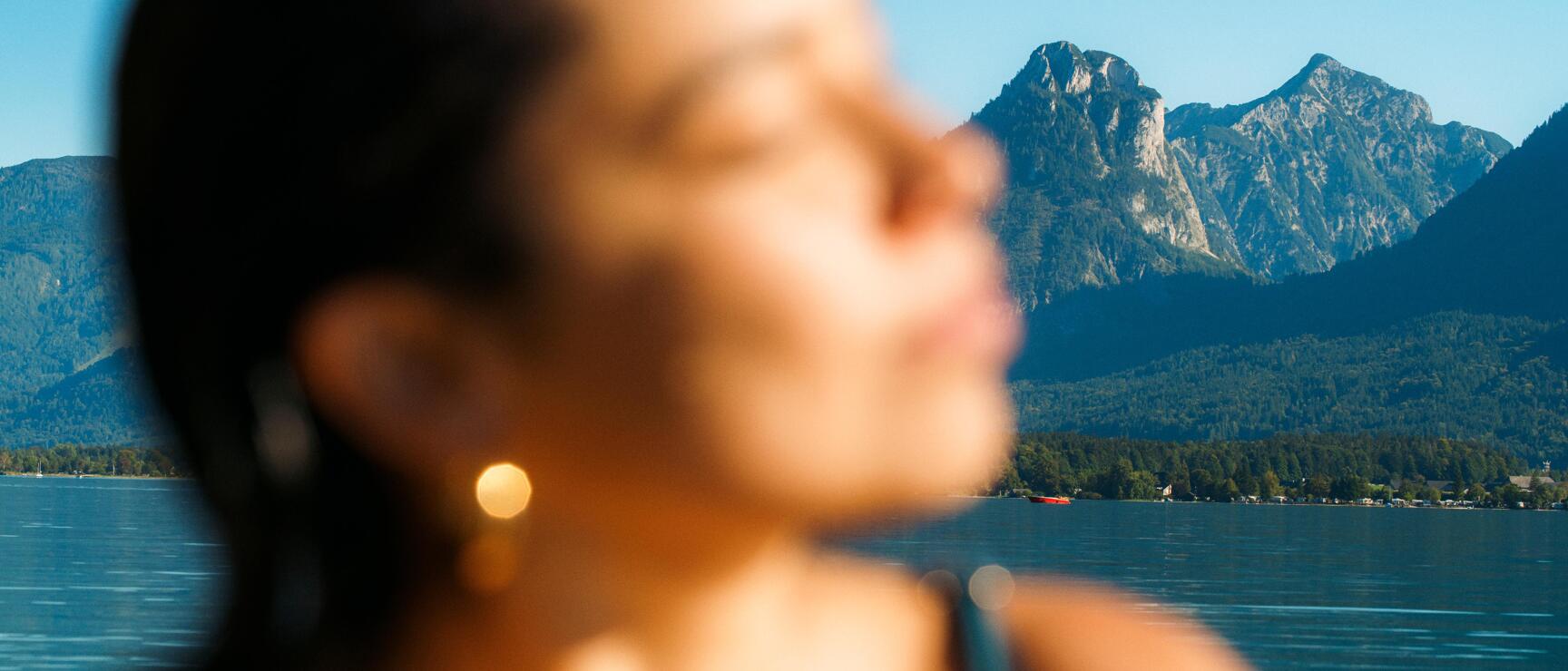 Woman in foreground (out of focus), blue lake and mountain peak beyond under clear sky.