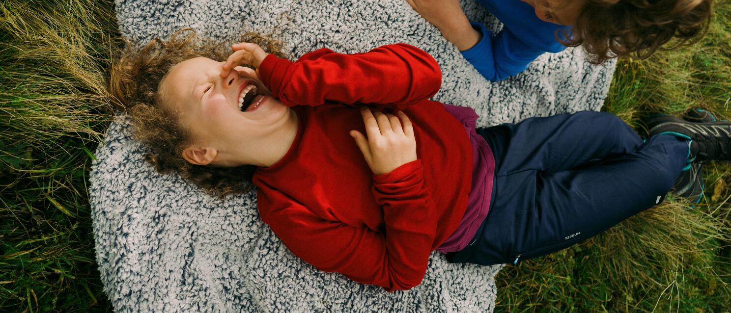 Two children laughing on a gray blanket in the grass, one in red, one in blue.