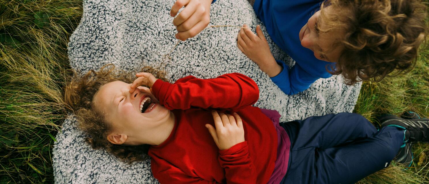 Zwei Kinder liegen lachend auf einer grauen Decke im Gras, eines in Rot, eines in Blau.