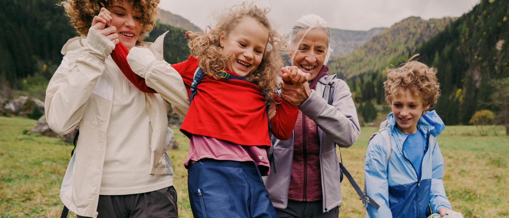 Two women swinging a laughing girl on a mountain meadow, boy beside them, forested mountains in the background.