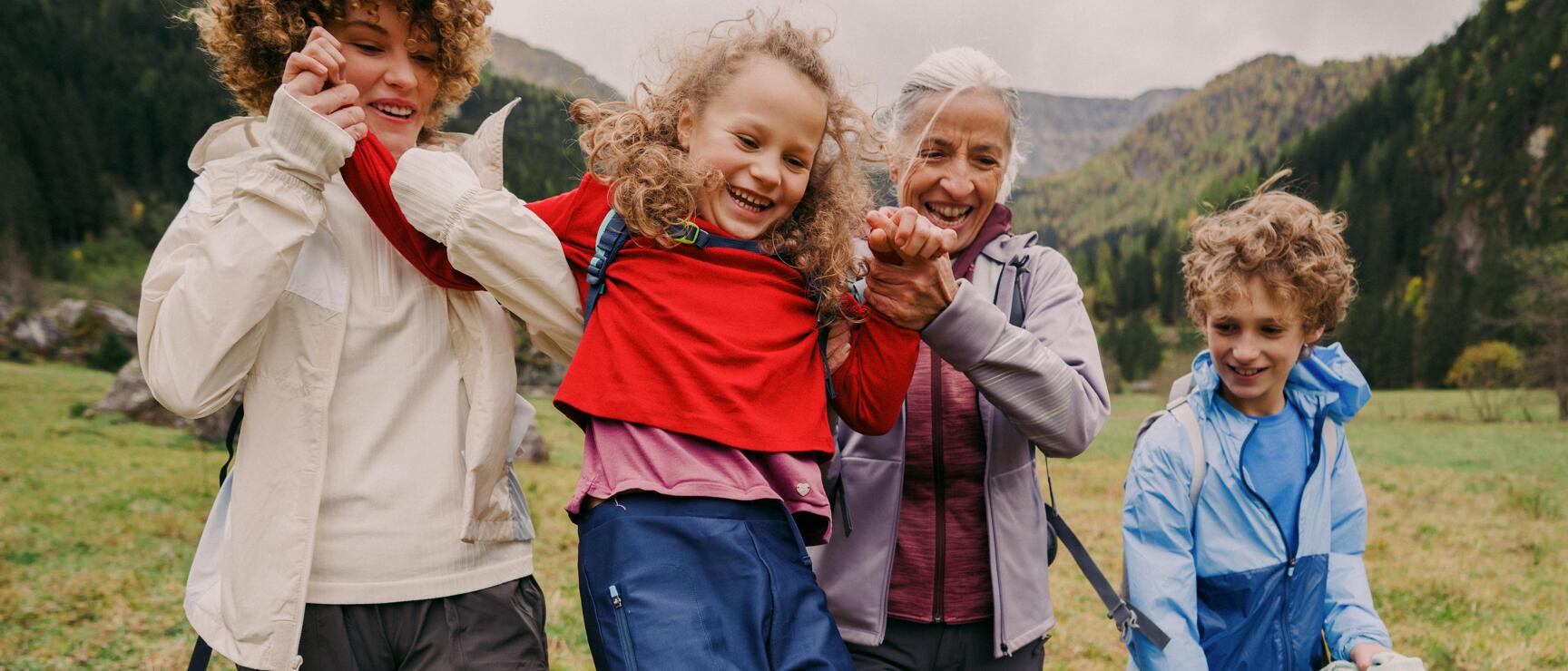 Two women swinging a laughing girl on a mountain meadow, boy beside them, forested mountains in the background.
