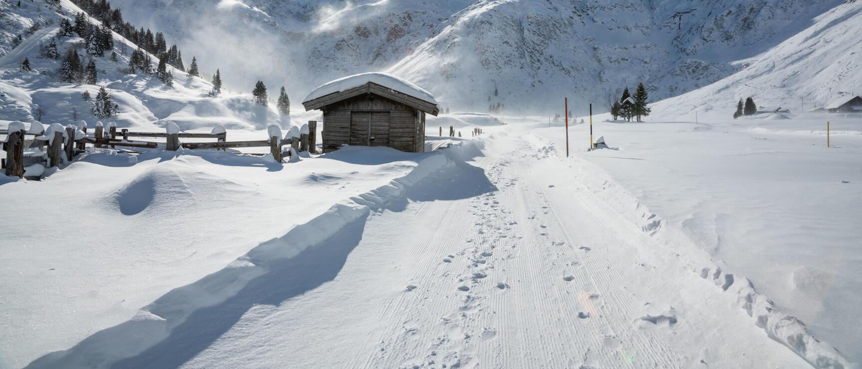 Snow-covered alpine hut beside a cross-country ski trail in a winter high valley with snow-capped mountain peaks in the background.
