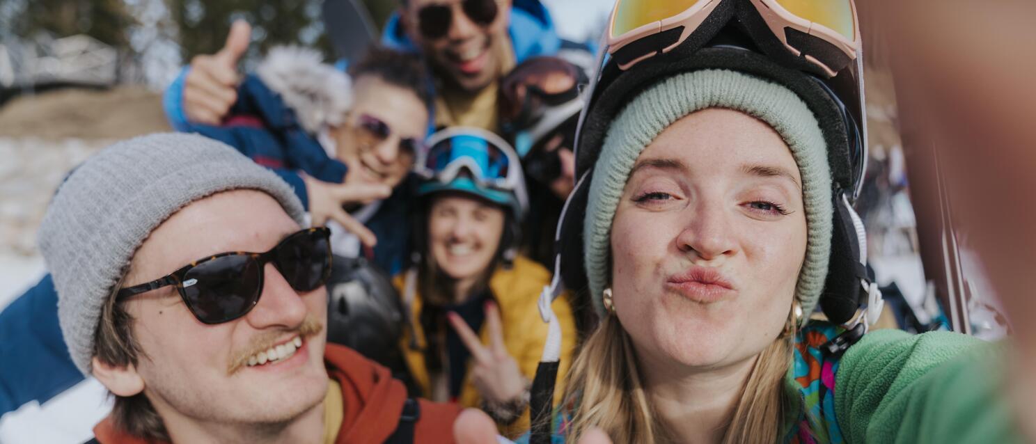 Gruppe junger Wintersportler macht Selfie im Schnee, mit Helmen, Skibrillen und bunter Winterkleidung.