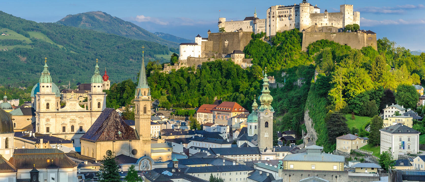 Salzburg old town with cathedral, church spires and Hohensalzburg Fortress on wooded cliffs, green mountains in background.