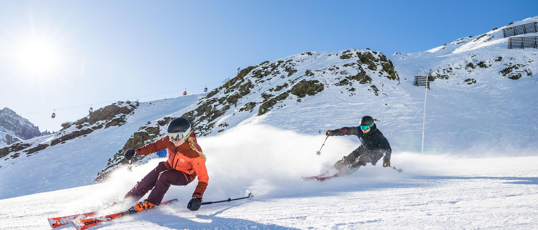Two skiers on sunny slope with snow spray clouds, cable car and rocky summit in background.