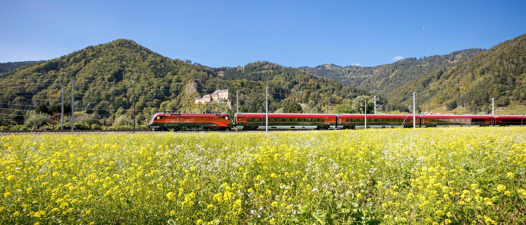 Red Railjet train passing yellow flower field with forested hills and castle in background.