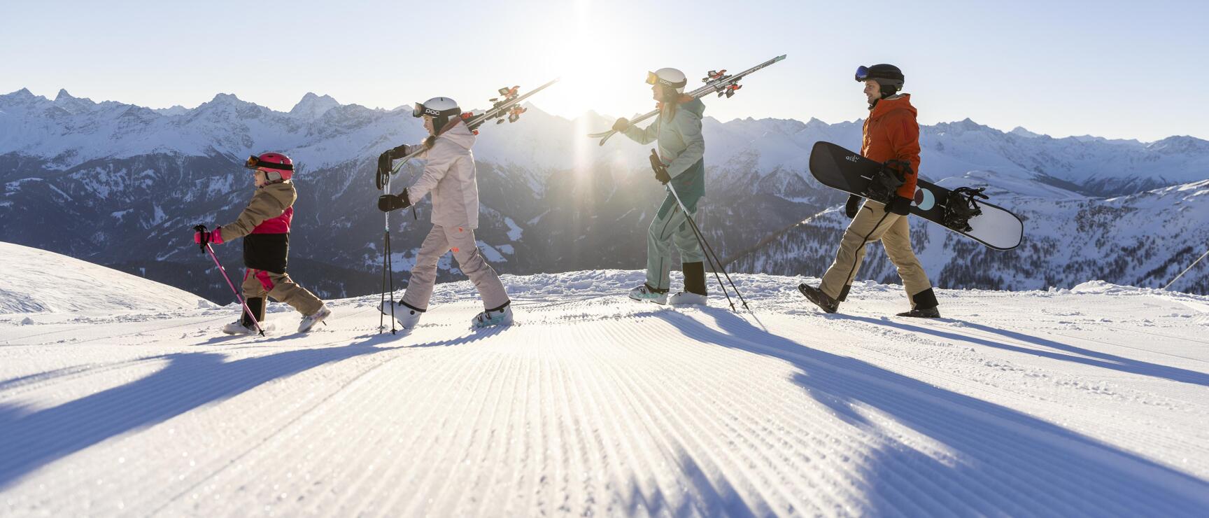 Vier Personen mit Skiern und Snowboard gehen auf präparierter Piste, schneebedeckte Alpen im Gegenlicht.