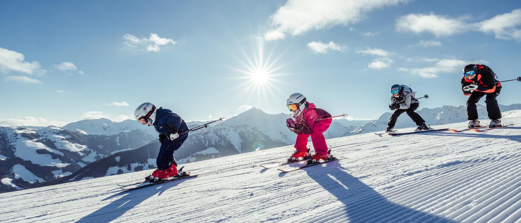 Four skiers on a prepared slope in sunshine, snow-covered mountains in the background.