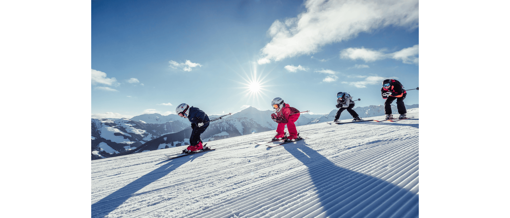 Four skiers on a prepared slope in sunshine, snow-covered mountains in the background.
