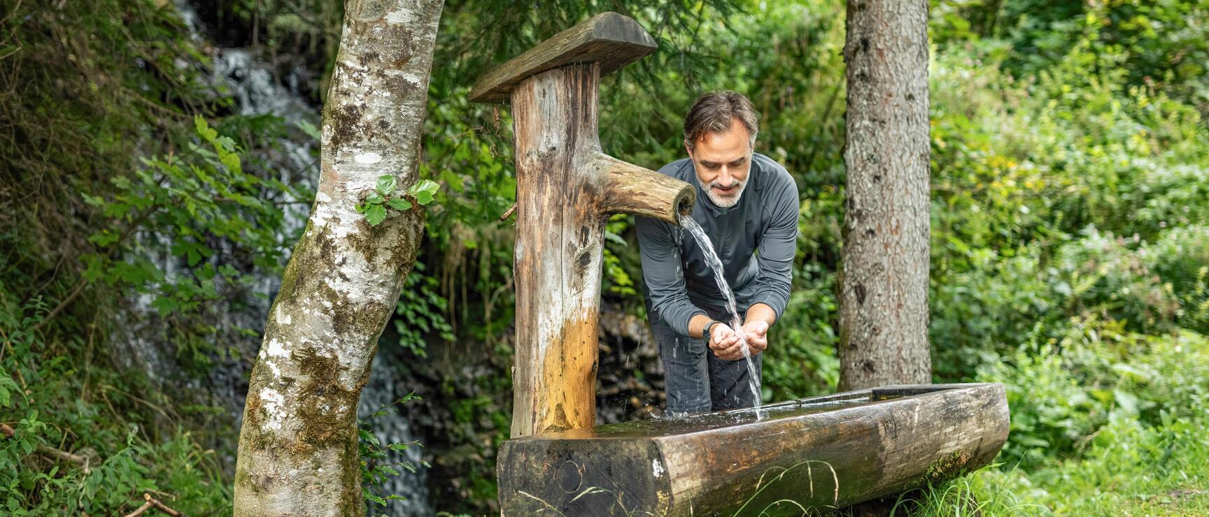 Mann erfrischt Hände an einem hölzernen Brunnen im Wald, Wasserfall im Hintergrund.