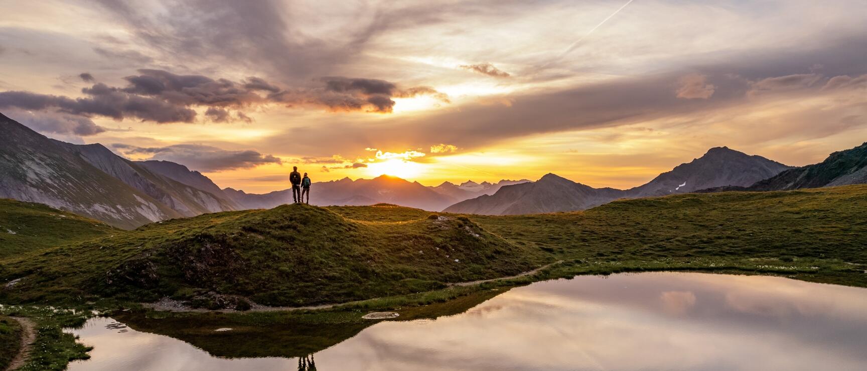 Two people as silhouettes on a hill at sunset, mountain lake with reflection, Alps in the background.
