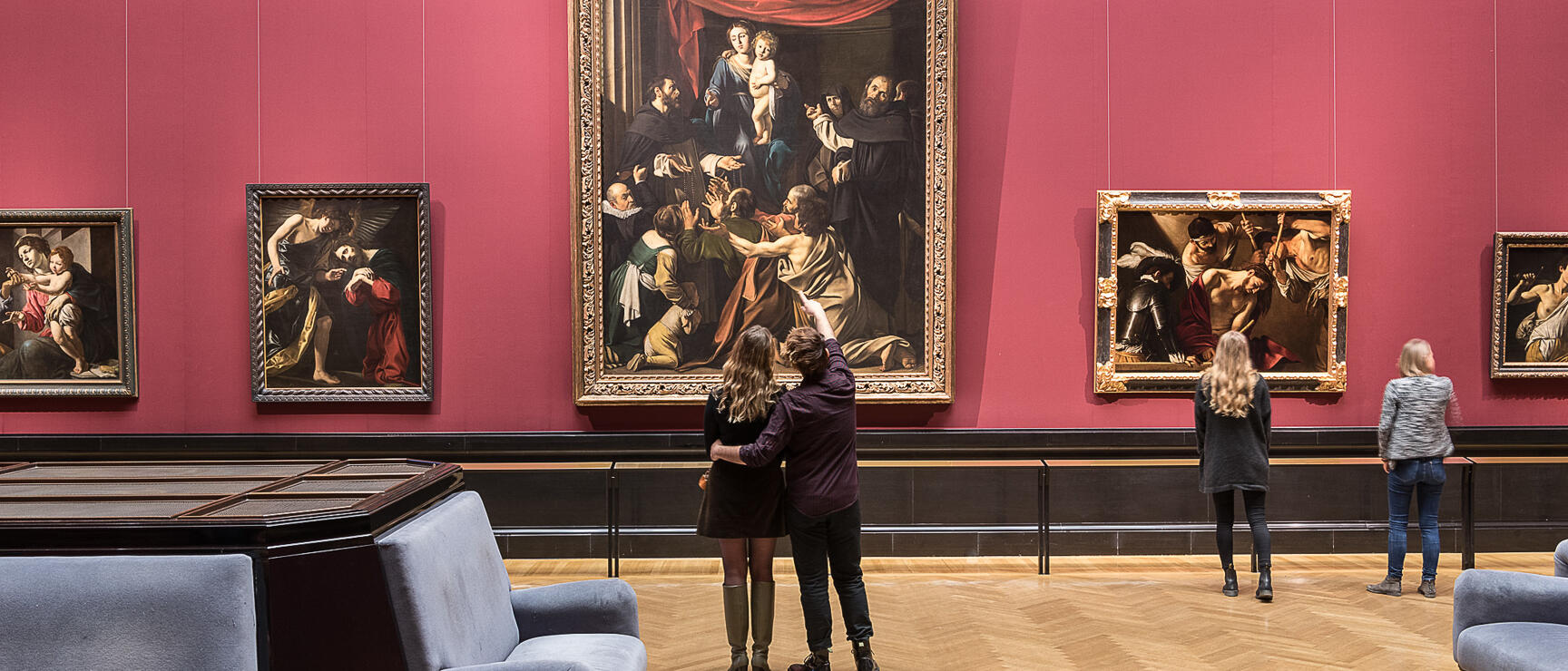 Museum room with red wall, four people viewing large old master paintings, parquet floor.