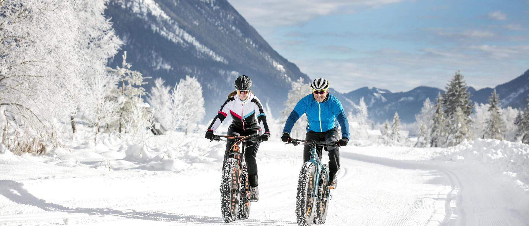 Two helmeted people riding fat bikes on a snow-covered trail with mountain panorama in the background.