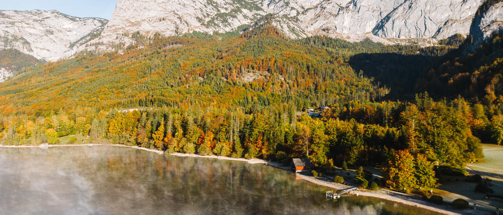 Aerial view of mountain lake in autumn, mixed colorful forest, boathouse on shore, rocky peak in background.