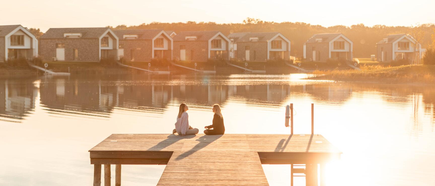 Two women sitting on a wooden dock by a calm lake, holiday cottages backlit in the background.