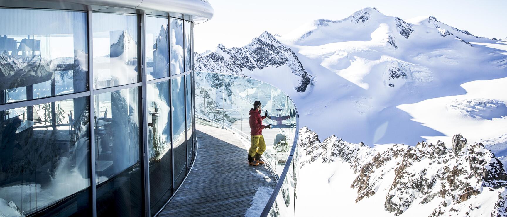 Person in red jacket on viewing platform with glass front, snow-capped peaks and glacier in the background.