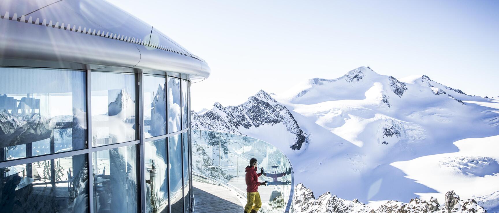 Person in roter Jacke auf Aussichtsplattform mit Glasfront, schneebedeckte Gipfel und Gletscher im Hintergrund.