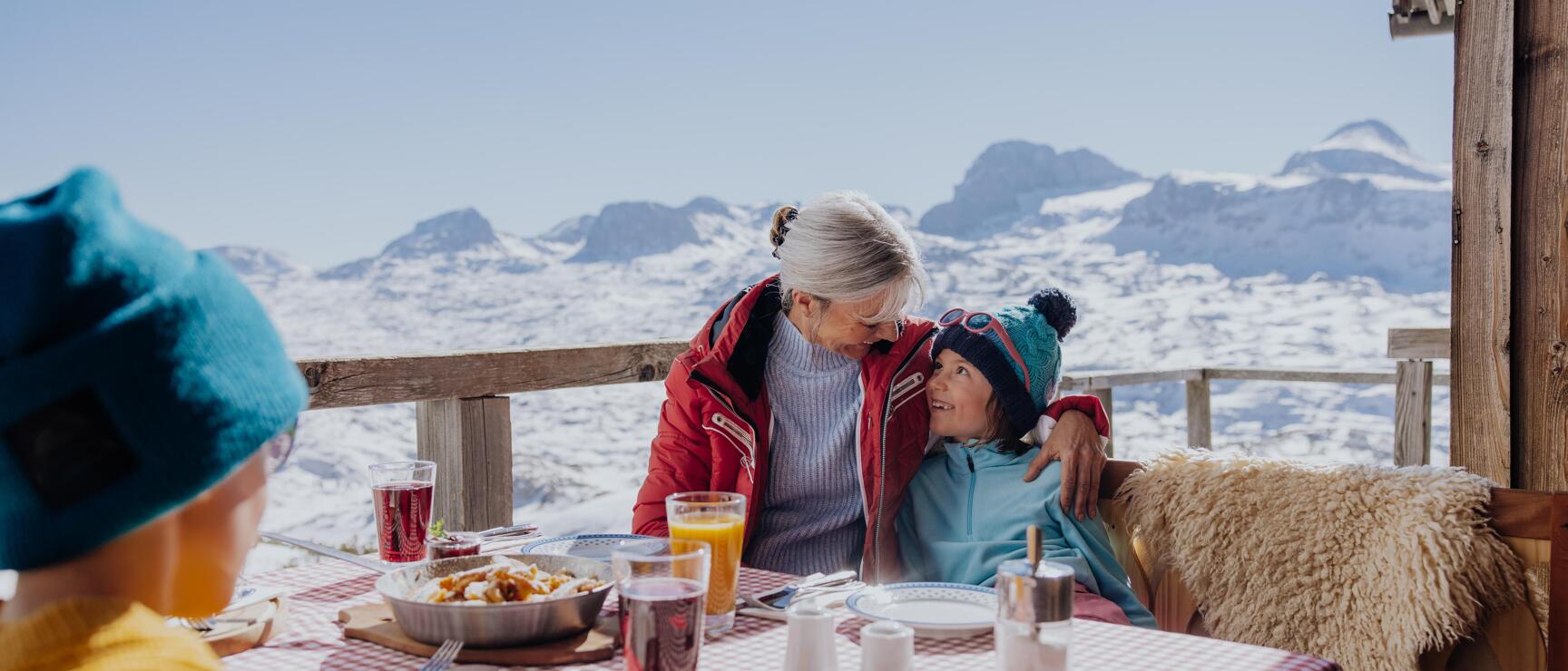 Woman and child sitting at a table on a snow-covered alpine hut terrace with snow-capped mountains in the background.