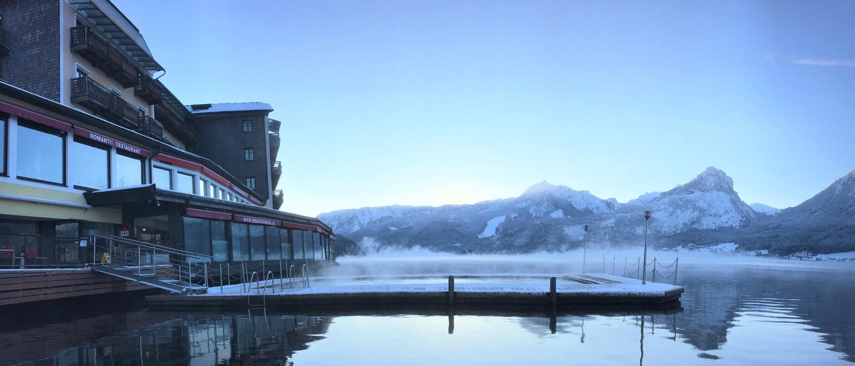 Snow-covered wooden jetty at a lake in winter, hotel with restaurant on the left, snow-capped mountains in the background.