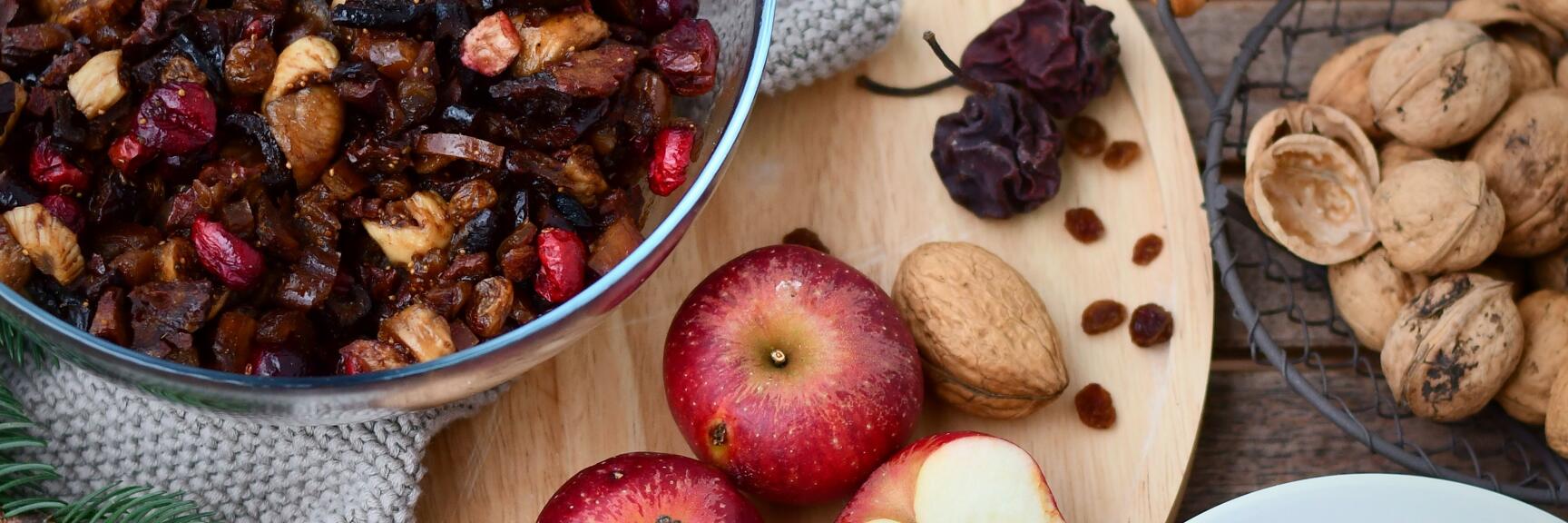 Glass bowl with dried fruit, red apples, nuts and raisins on wooden board, fir branches as decoration.