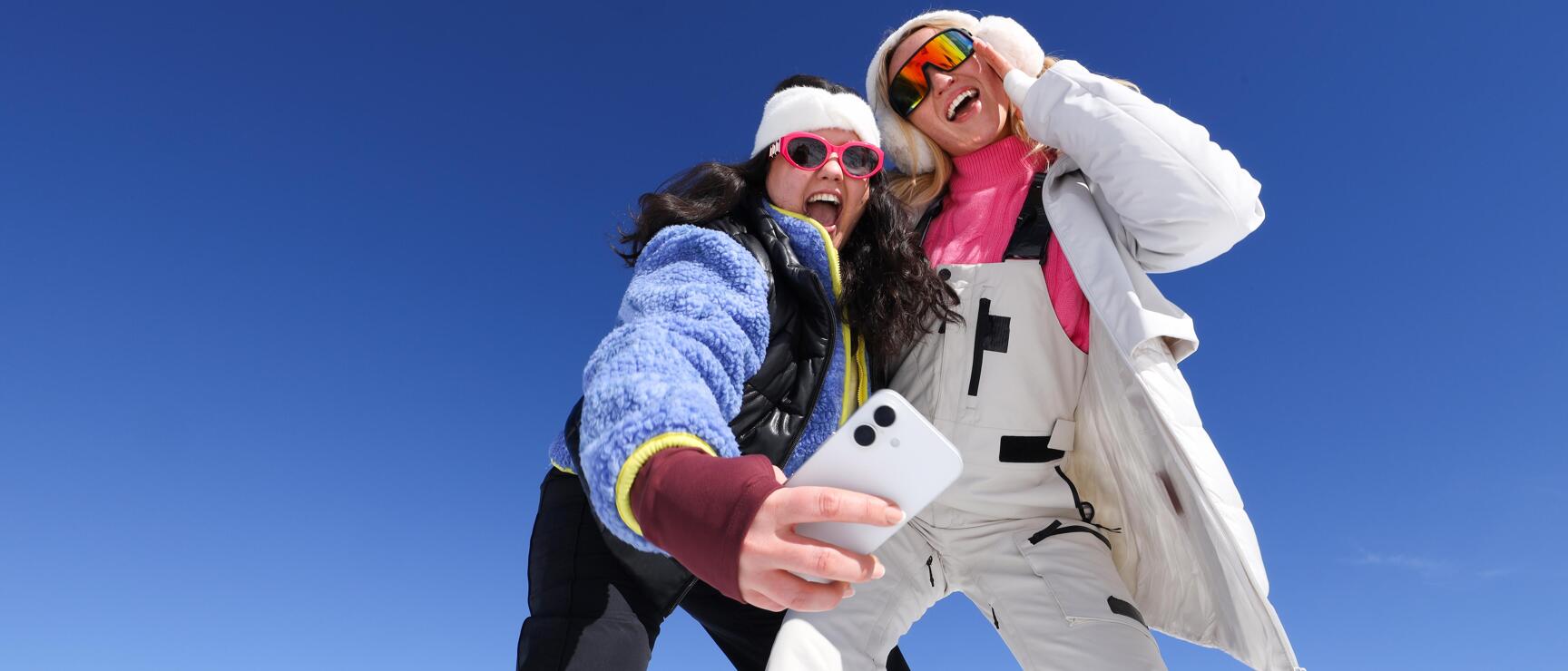 Two laughing women in ski outfits taking a selfie on a snow-covered mountain peak, blue sky.
