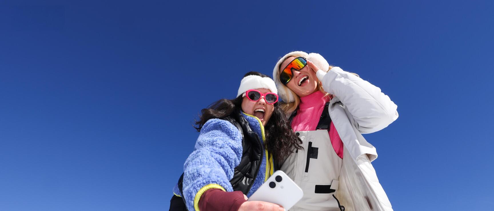 Two laughing women in ski outfits taking a selfie on a snow-covered mountain peak, blue sky.