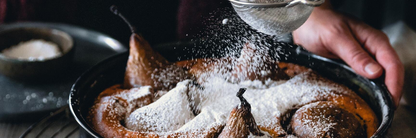 Hands dusting pear cake in springform with icing sugar through a sieve.
