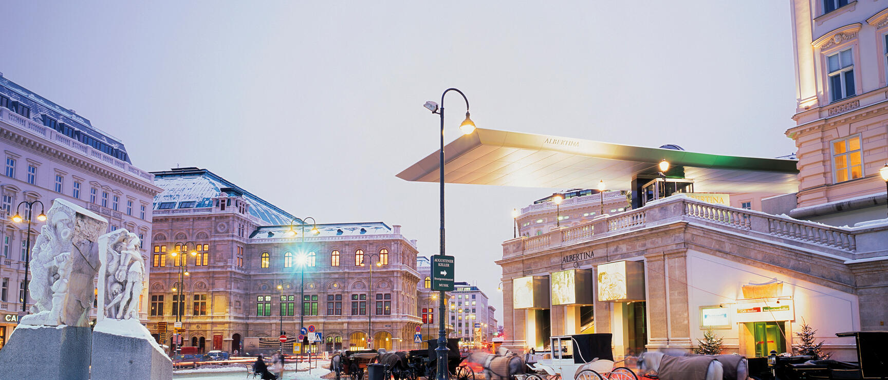 Verschneiter Albertinaplatz Wien bei Abenddämmerung, Fiaker, Staatsoper und beleuchtete Albertina im Hintergrund.