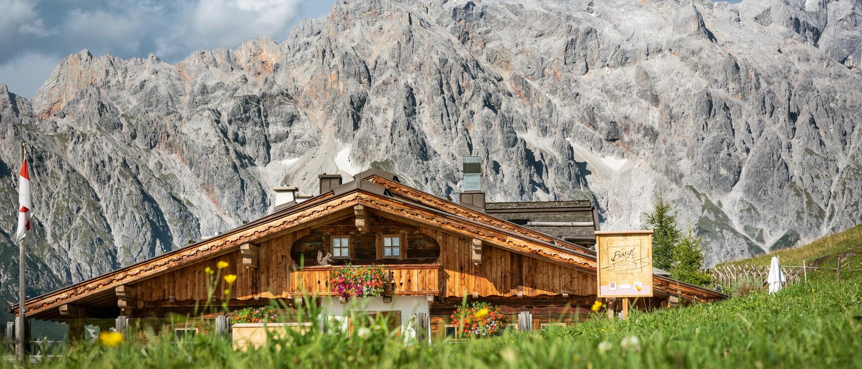 Wooden alpine hut with flower boxes on green mountain meadow, rocky mountain range in background, Austrian flag on left.