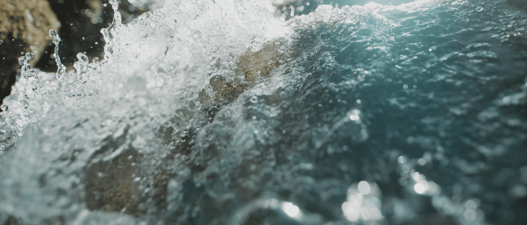Close-up: Water splashing over wet rocks, blue-turquoise whitewater backlit.