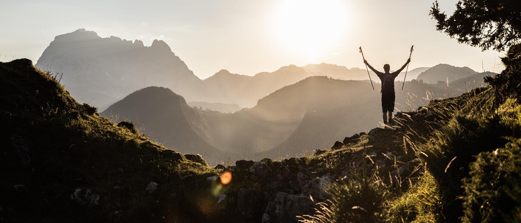 Hiker with raised trekking poles on mountain ridge, silhouette against backlighting, mountain panorama in background.