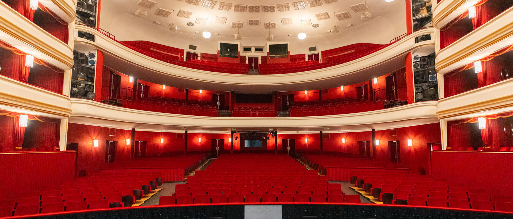 Empty theatre hall with red seating rows, two balconies and orchestra pit furnishings in the foreground.