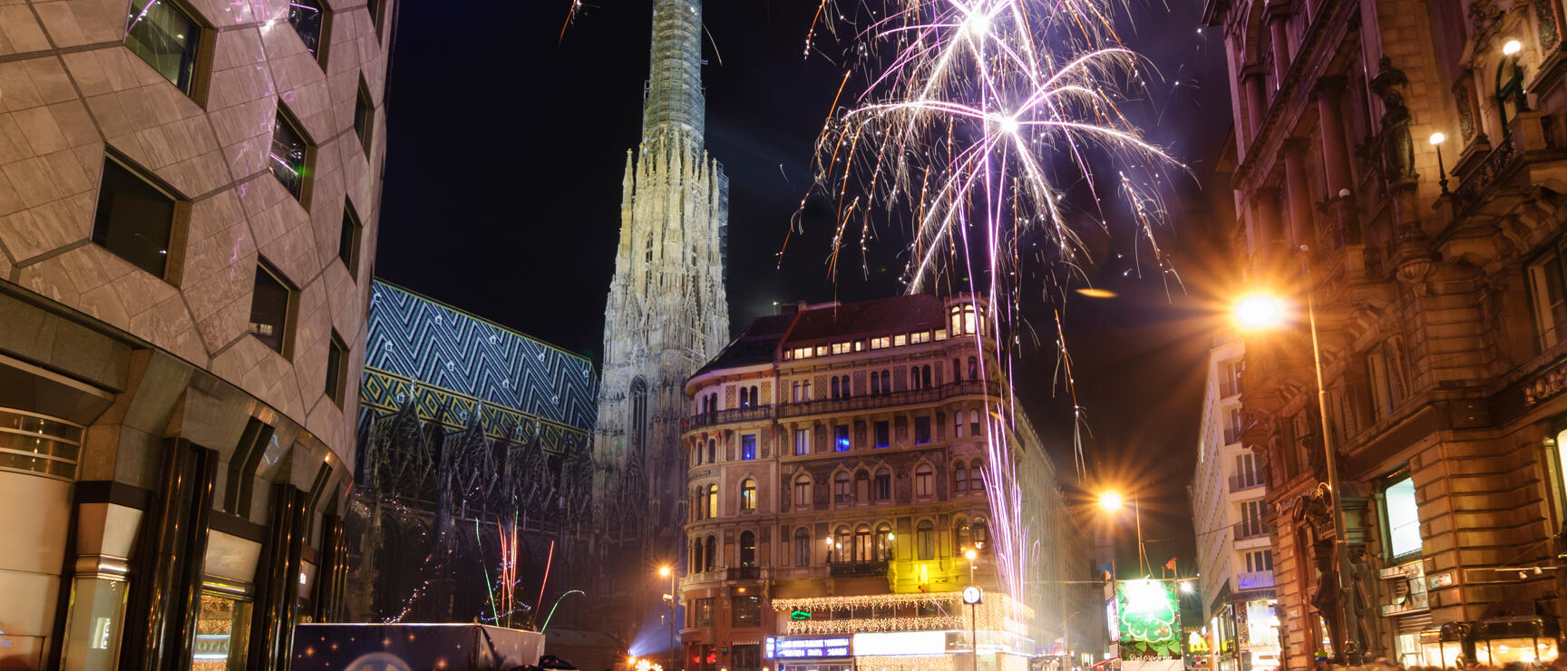 Fireworks over St. Stephen's Cathedral in Vienna at night, crowd of people on Kärntner Street in foreground.