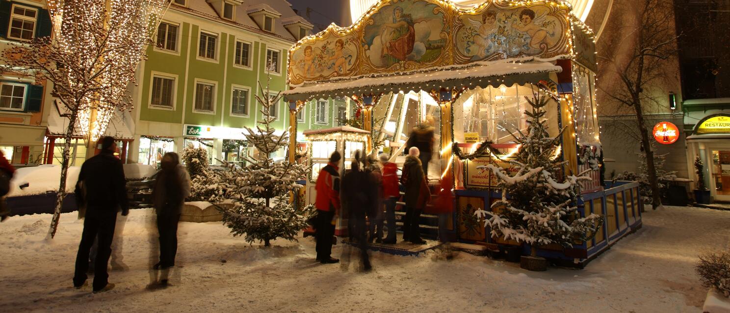 Christmas market at night with illuminated Ferris wheel, carousel and snow-covered fir trees, people in the square