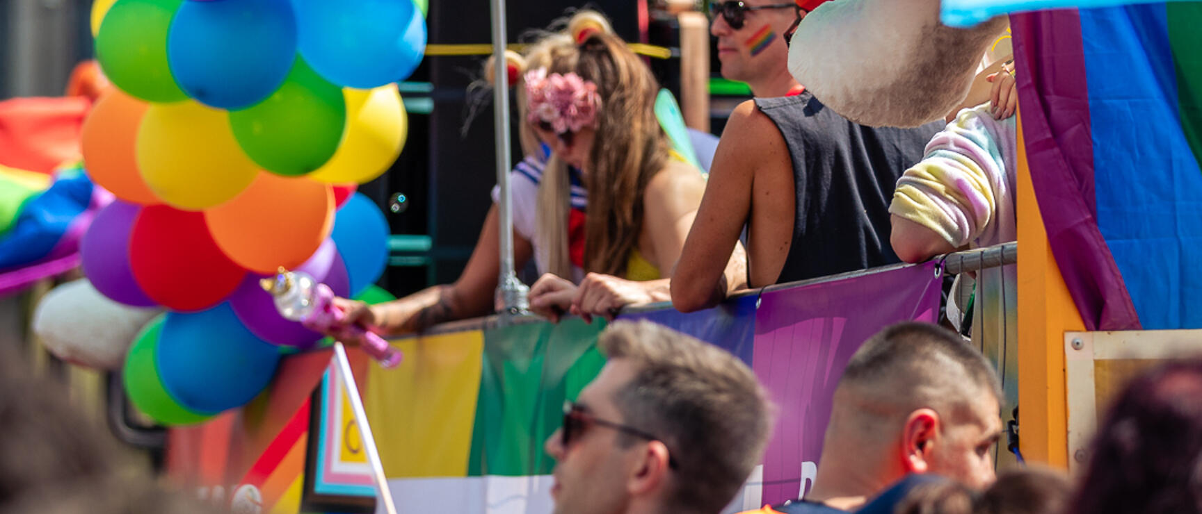 Festive float with colourful balloon arch and crowd, rainbow and trans flags, Gründerzeit building facade in background.