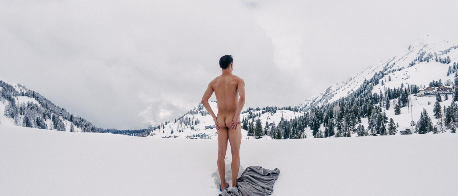 Nackter Mann von hinten in verschneiter Berglandschaft, graues Tuch im Schnee, bewaldete Hänge im Hintergrund.