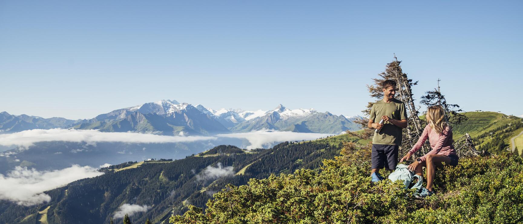 Two hikers resting on mountain ridge with snow-covered peaks and sea of clouds in background.