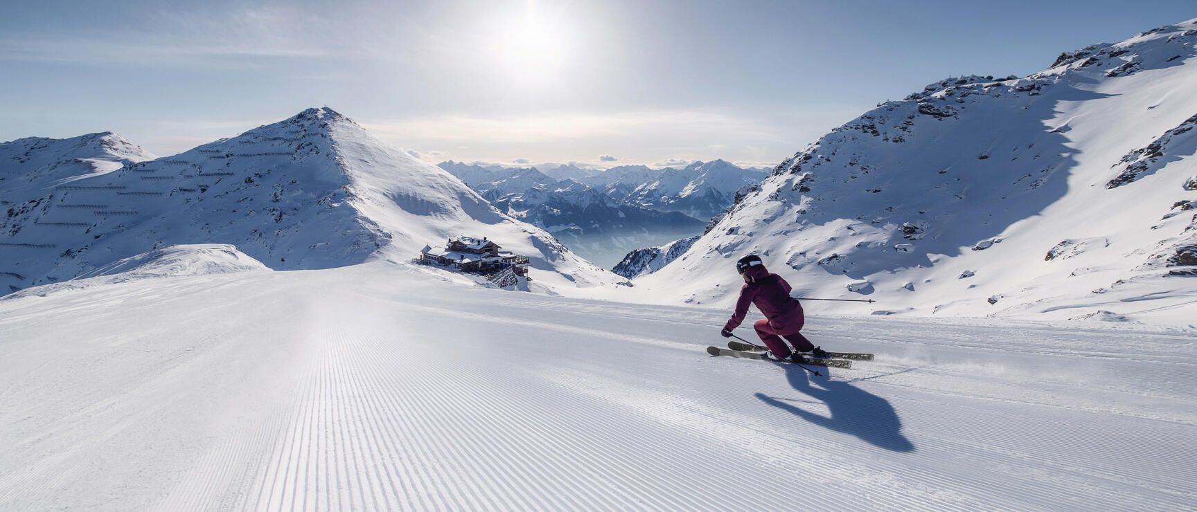 Skier in pink suit on prepared ski slope with mountain lodge and snow-covered peaks in background