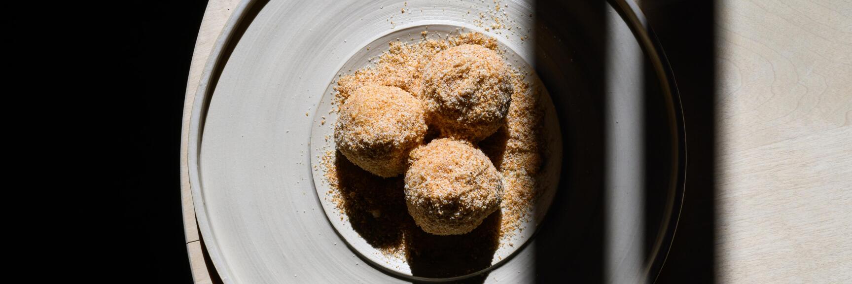 Drei kugelförmige, mit Sig – einer Bregenzerwälder Schokolade – gefüllte Knödel mit brauner Bröselpanade, in einem Teller mit Streuseln angerichtet, von oben fotografiert.