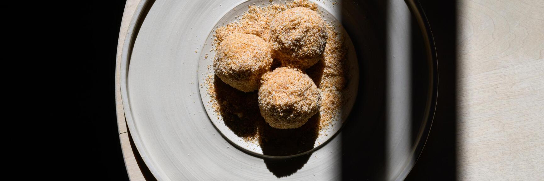Three spherical dumplings filled with Sig—a chocolate from the Bregenzerwald region—and coated in brown breadcrumbs, arranged on a plate with crumble topping, photographed from above.