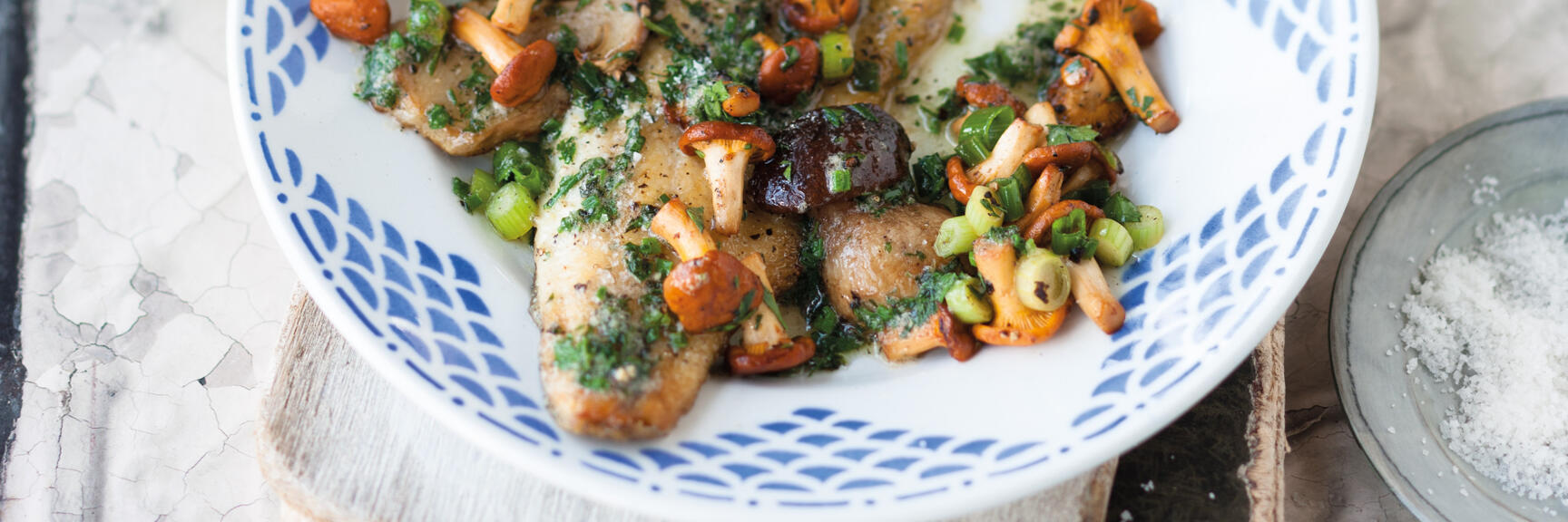 Gebratene Fischfilets mit Eierschwammerln, Kräutern und Frühlingszwiebeln auf blau-weißem Teller.