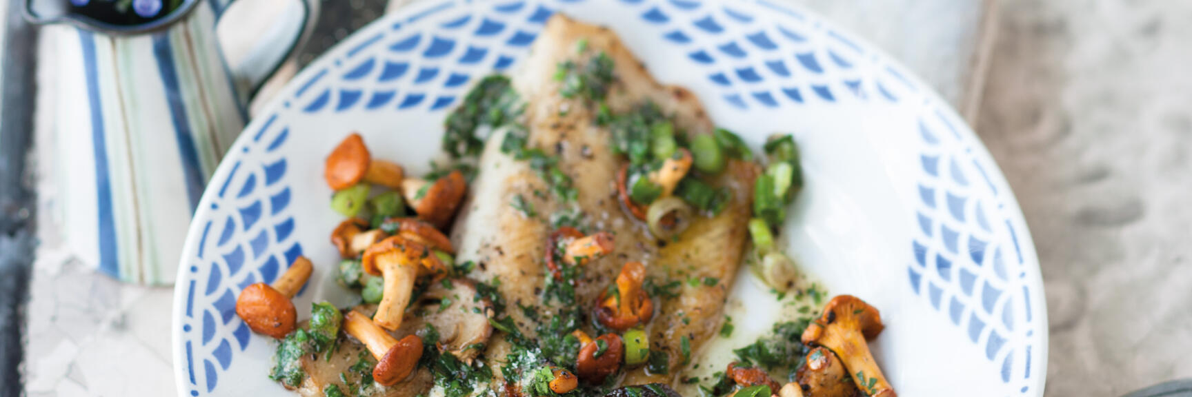 Fried fish fillets with chanterelles, herbs and spring onions on blue-white plate.