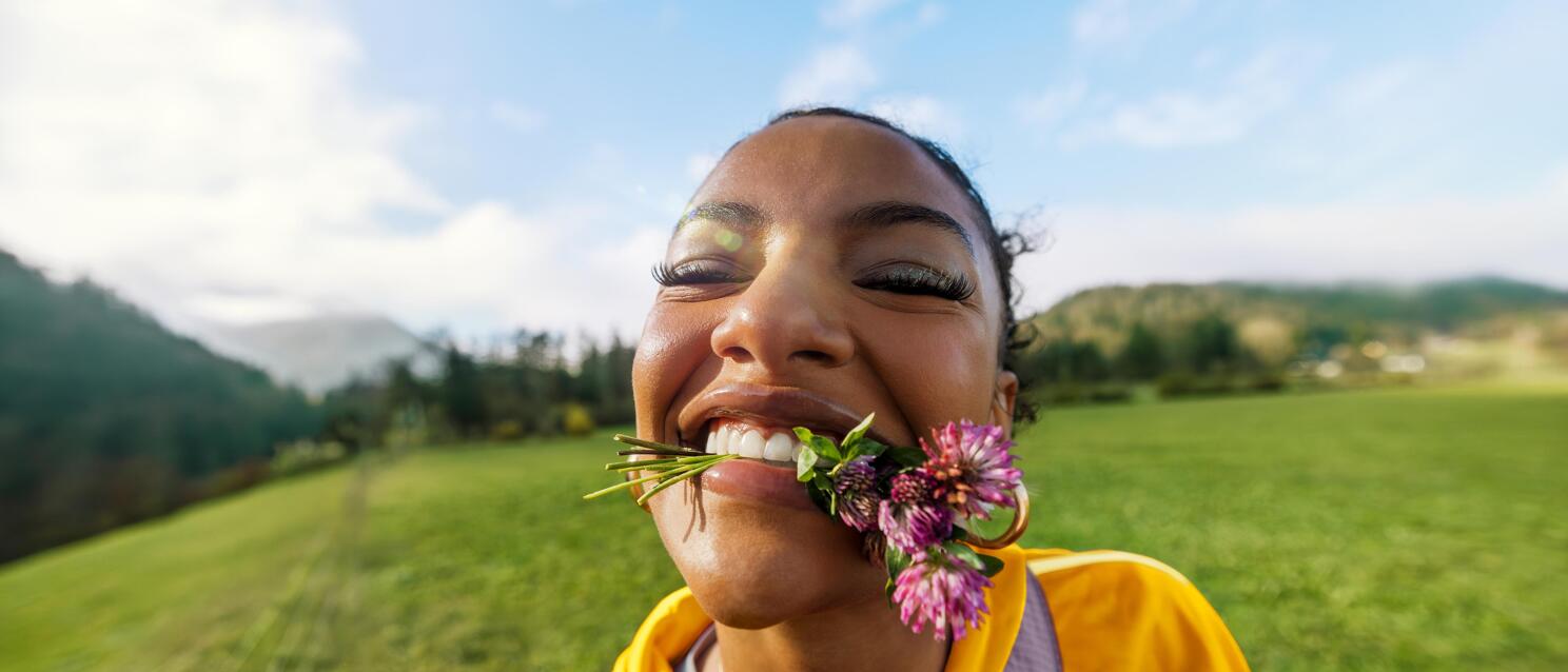 Smiling woman with clover flowers in her mouth, yellow jacket, green mountain meadow and blue sky in the background.