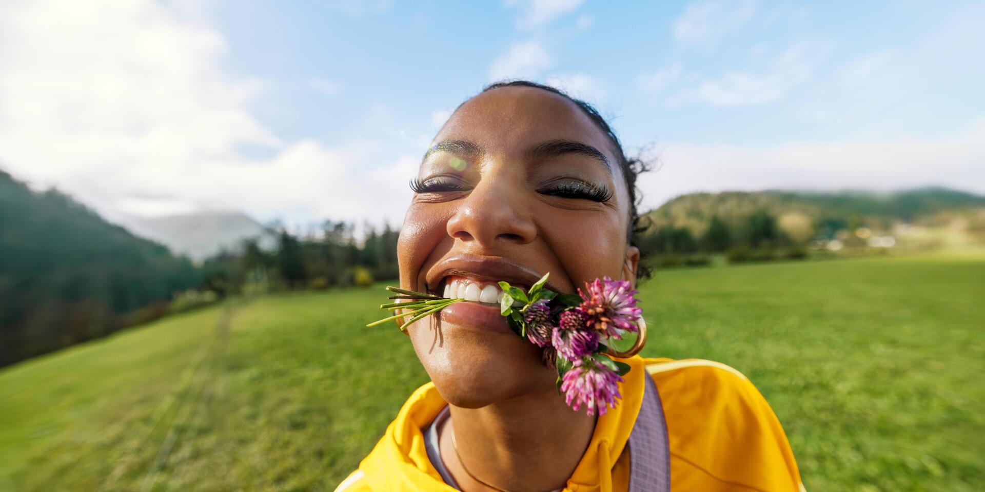 Lachende Frau mit Kleeblüten im Mund, gelbe Jacke, grüne Bergwiese und blauer Himmel im Hintergrund.