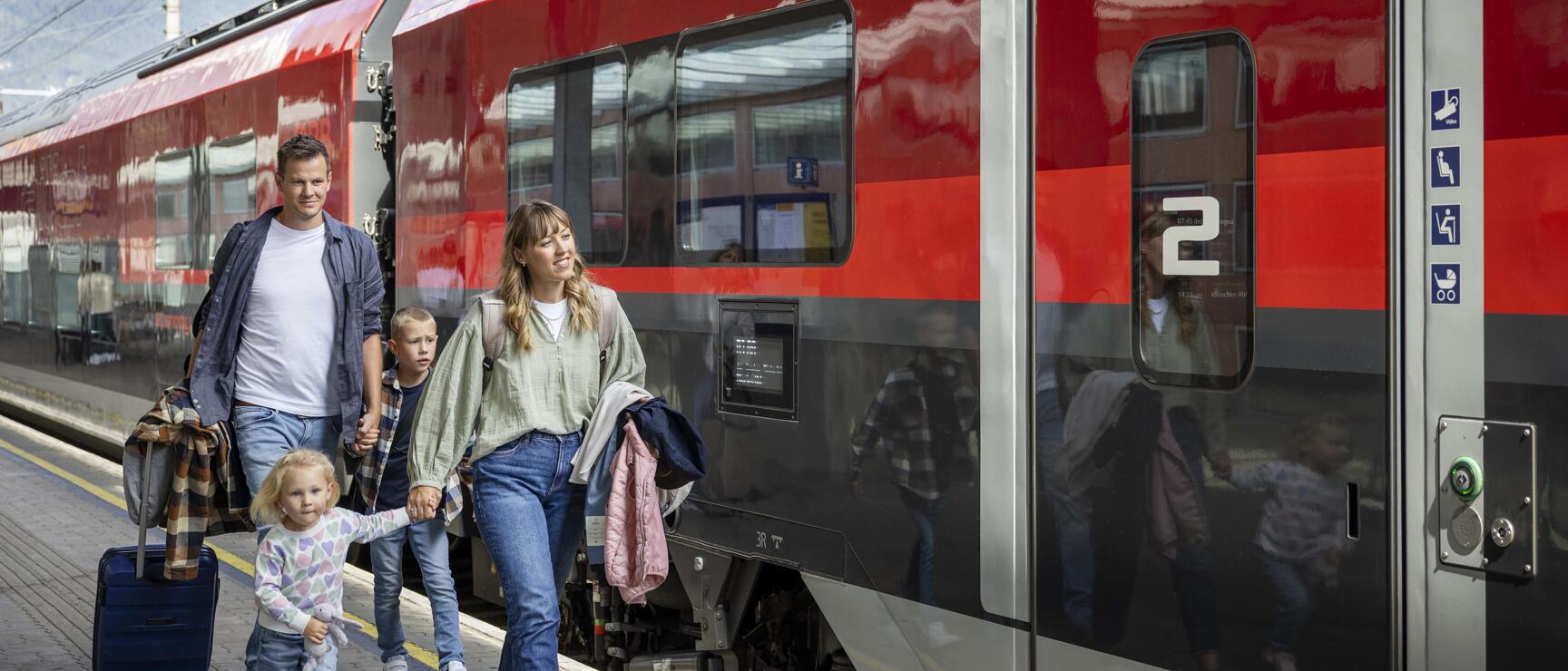 Two adults and two children with suitcases on platform beside red ÖBB train, mountains in background.