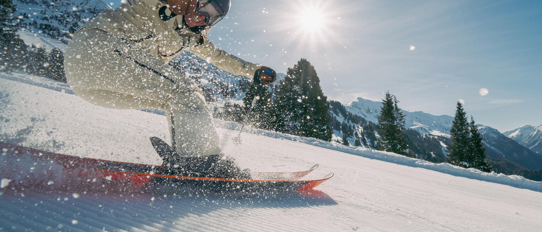 Skier with helmet on prepared slope, snow spray, fir trees and snow-covered mountains in sunshine.