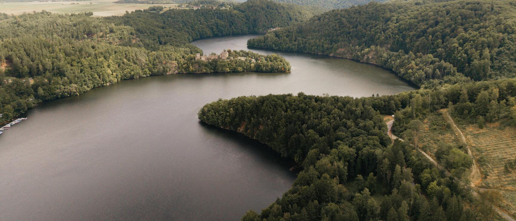 Aerial view of a reservoir surrounded by forested hills with a small castle ruin on the shoreline.