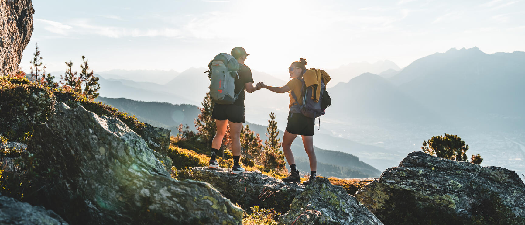 Two hikers with backpacks on rocks, holding hands, Alpine panorama backlit.