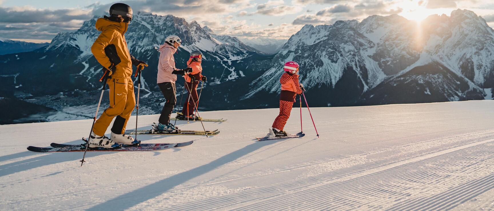 Four skiers on groomed slope with snow-covered mountain range and sun in background.